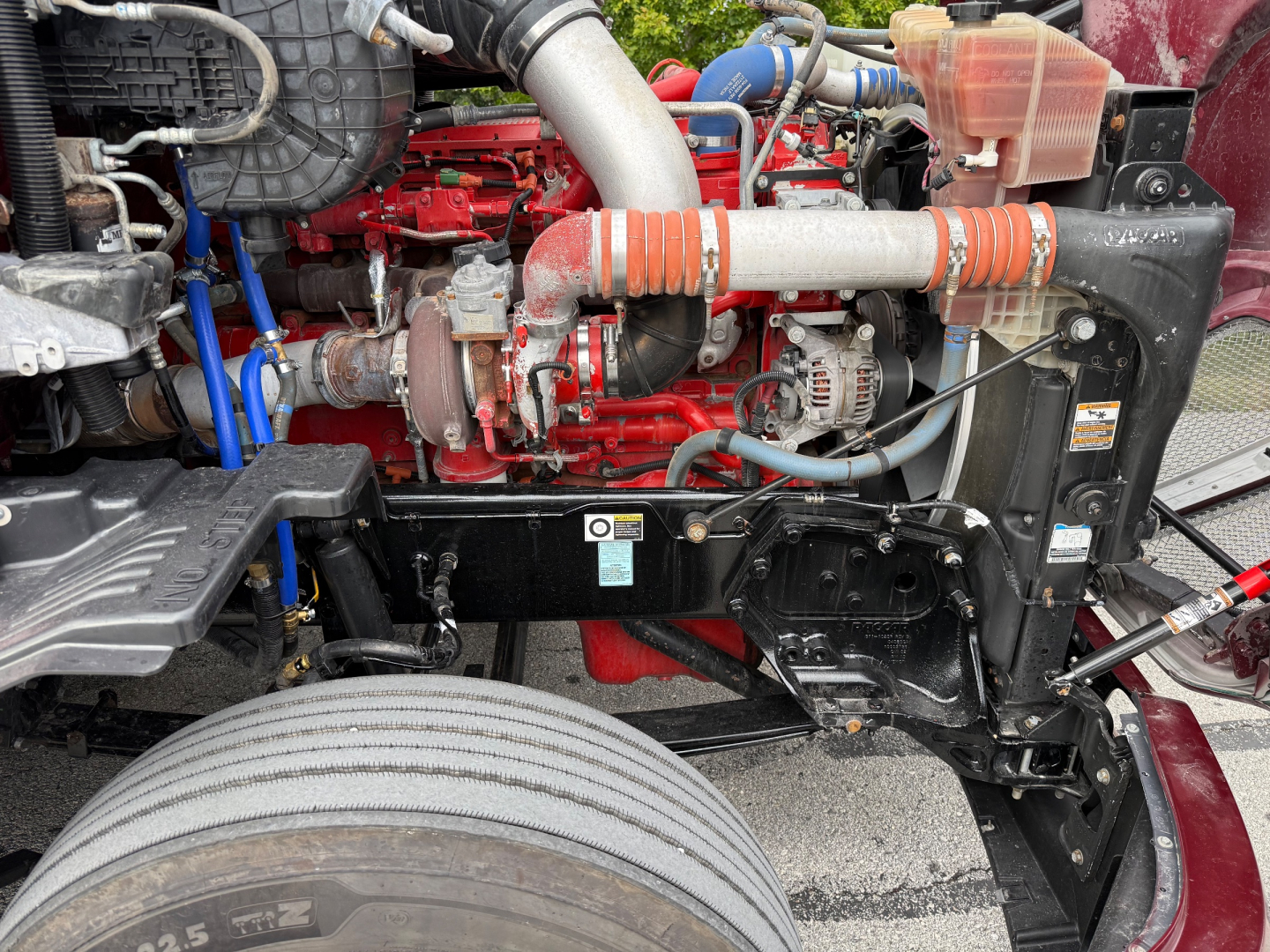 Kenworth T680 engine bay showing red engine, hoses, and coolant reservoir with orange braided hoses and black radiator support.