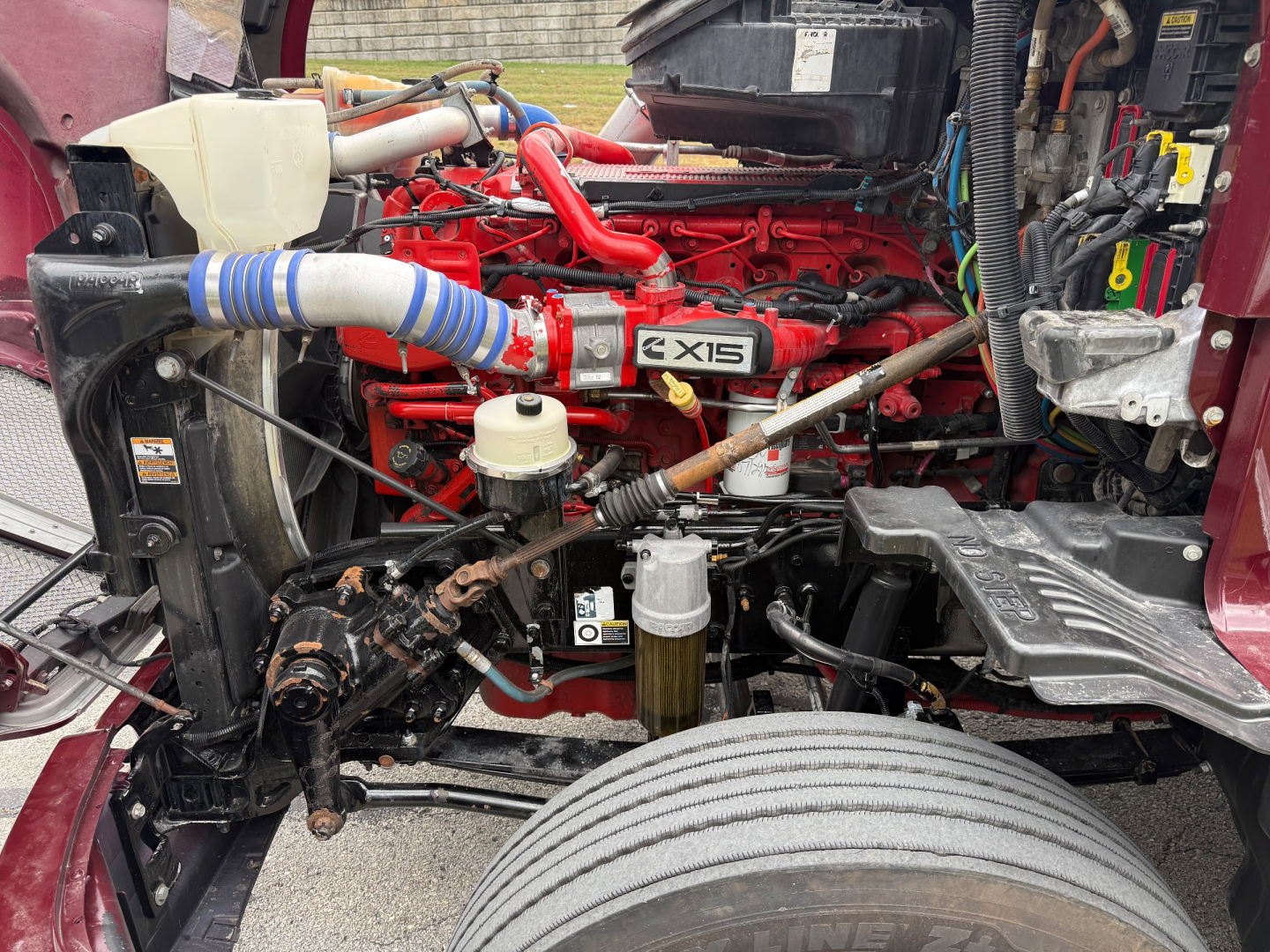 Kenworth T680 engine bay with red engine, X15 badge, white coolant reservoir, radiator, hoses, and front wheel visible.