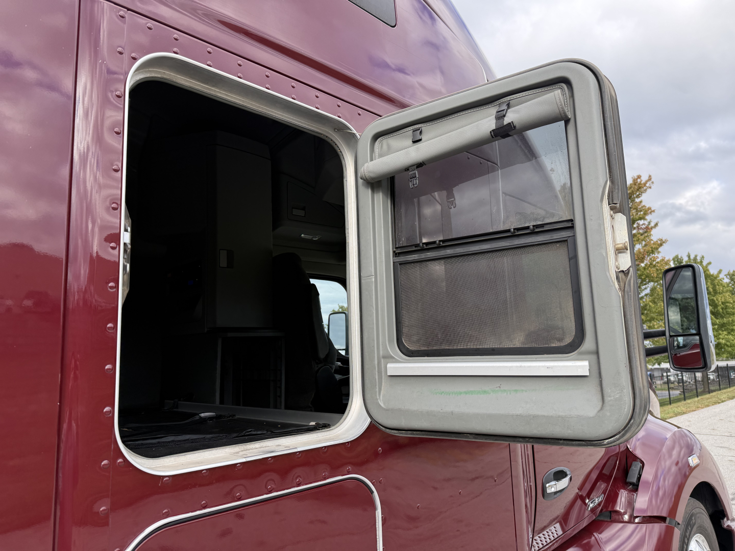 Kenworth T680 sleeper cab with open side window and door, maroon exterior, empty cargo area visible through window, roadside background.