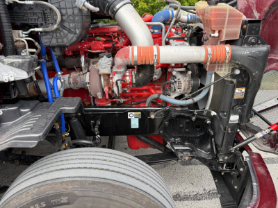 Kenworth T680 engine bay showing red engine, hoses, and coolant reservoir with orange braided hoses and black radiator support.