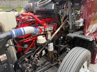 Kenworth T680 engine bay with red engine block and coolant hoses visible, air intake piping, hydraulic lines, and front wheel well; X15 badge on engine.