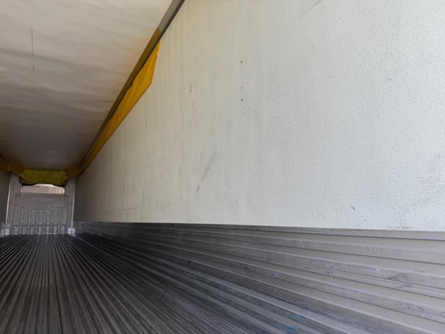 Trailer interior showing ribbed metal floor and white side wall, with a yellow tarpaulin strip along the top edge.