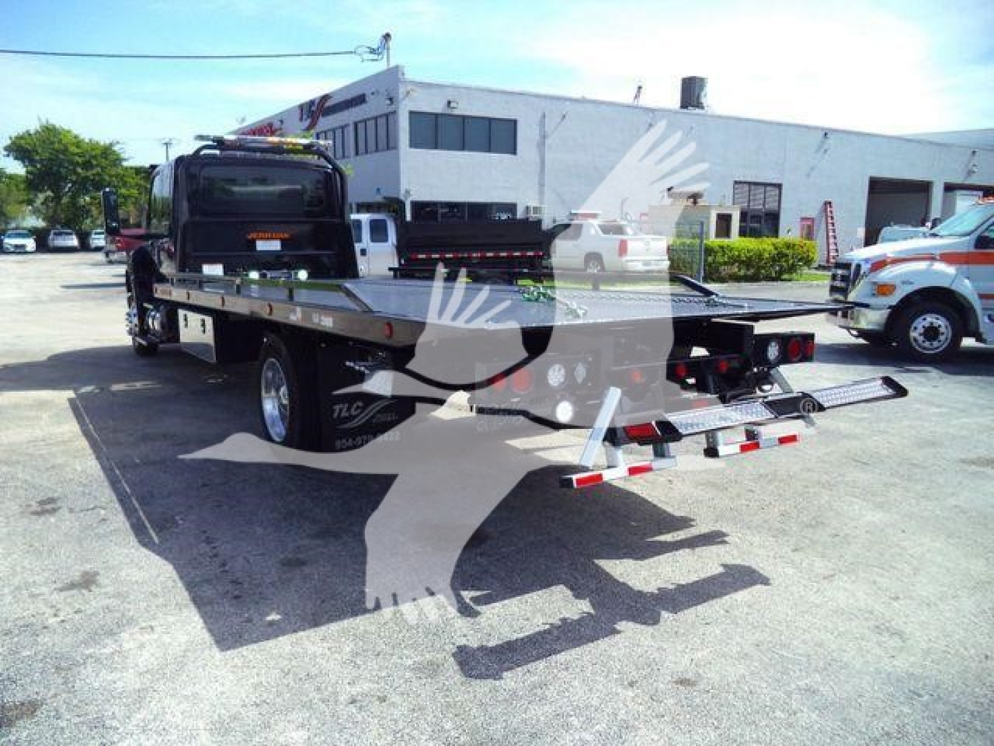Flatbed tow truck in a dealership lot with watermark, showing a flatbed with wheel ramps and rear lights.
