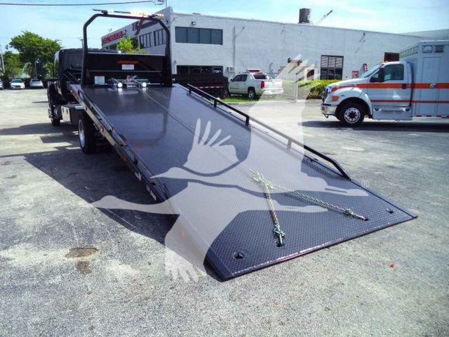 Flatbed tow truck from rear-left perspective with a long black steel ramp bed, metal chains, and a high side railing; a white building and ambulances visible in background.