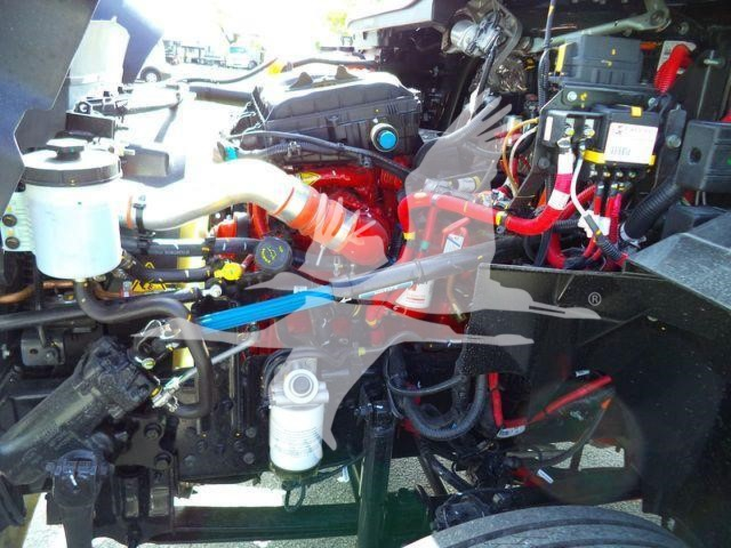 Truck engine bay with label text obscured; multiple hoses, battery, and coolant reservoir visible inside a heavy-use diesel engine compartment.