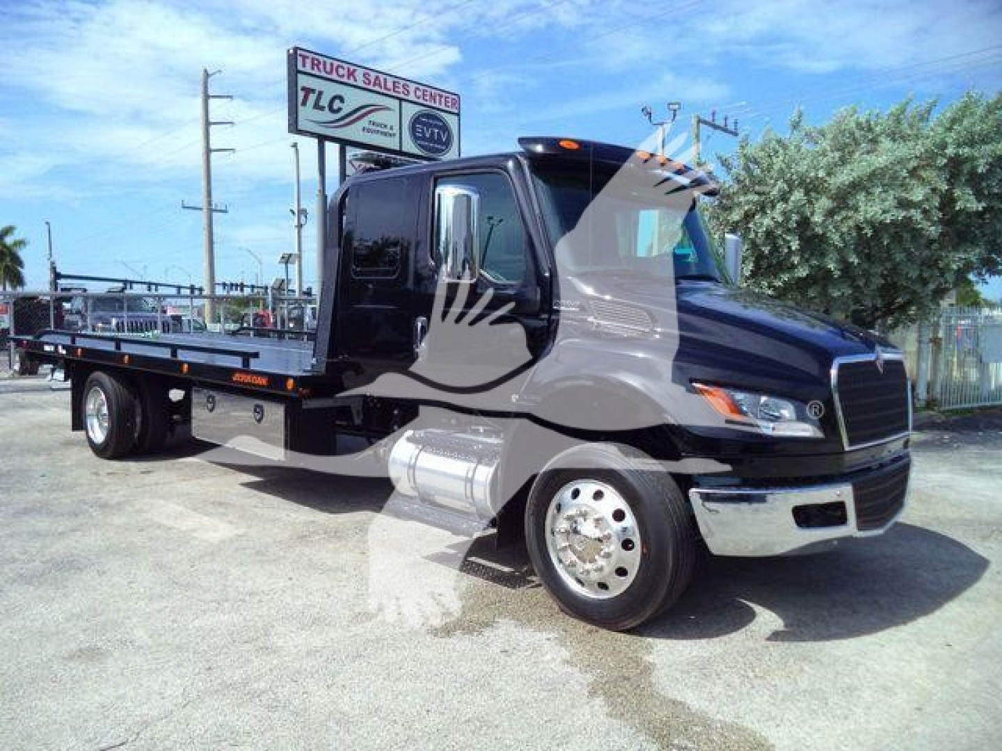 Tow truck flatbed with black cab, chrome bumper, and silver tank visible; sign in background reads Truck Sales Center TLC; no other legible labels on the truck.