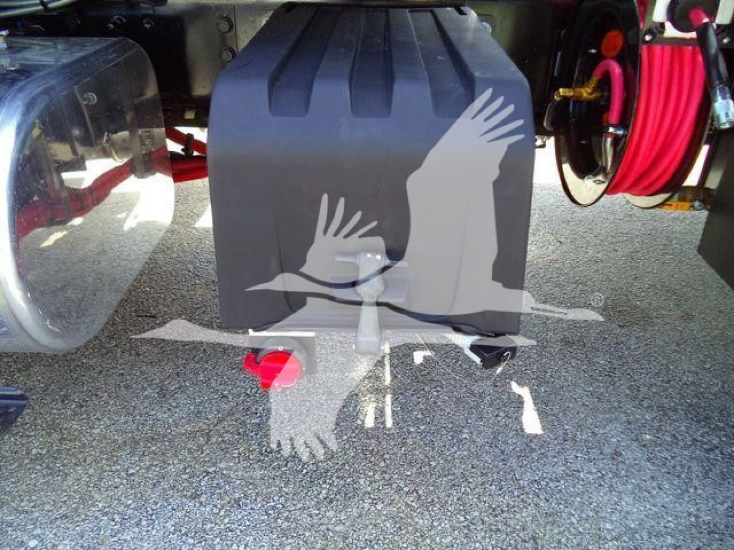 Tractor-trailer rear view showing black fuel/auxiliary tank with red tow eye and hooks, hoses coiled on right, and reflection of birds; MV607EXCABBLK stock number tag not visible.