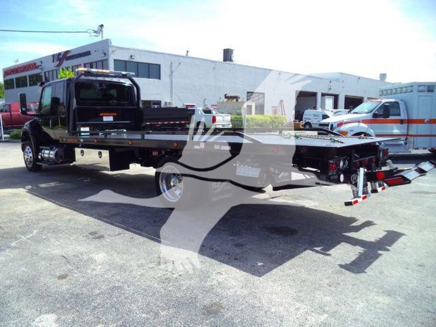 Truck hauler flatbed rollback with crane and wheel lift in foreground; International MV towing truck in background; dealership lot with buildings.