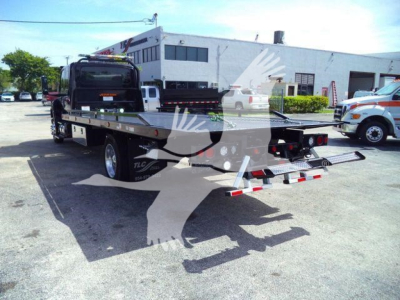 Flatbed tow truck in a dealership lot with watermark, showing a flatbed with wheel ramps and rear lights.