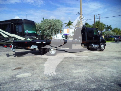 Flatbed rollback tow truck attached to a black International MV truck, side view with blue sky and palm trees.