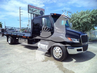 Tow truck flatbed with black cab, chrome bumper, and silver tank visible; sign in background reads Truck Sales Center TLC; no other legible labels on the truck.