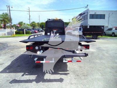 Rear view of a black flatbed rollback tow truck with warning lights and metal ramps, International MV chassis, in a dealership lot.