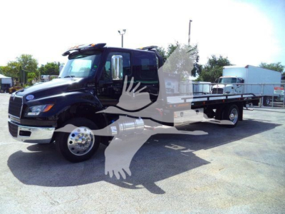 Black International MV truck with flatbed rollback body in a dealership lot; side view showing cab, mirrors, front grille, and long flatbed intake.