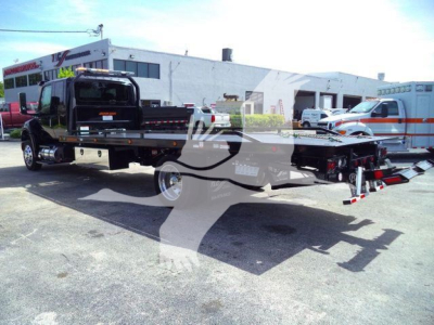 Truck hauler flatbed rollback with crane and wheel lift in foreground; International MV towing truck in background; dealership lot with buildings.