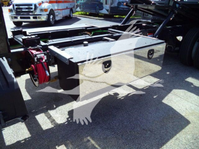 Truck equipment: a large aluminum tool/storage box mounted under a truck frame with a red reel hose or cable guide on left; flatbed structure and wheels visible in background.