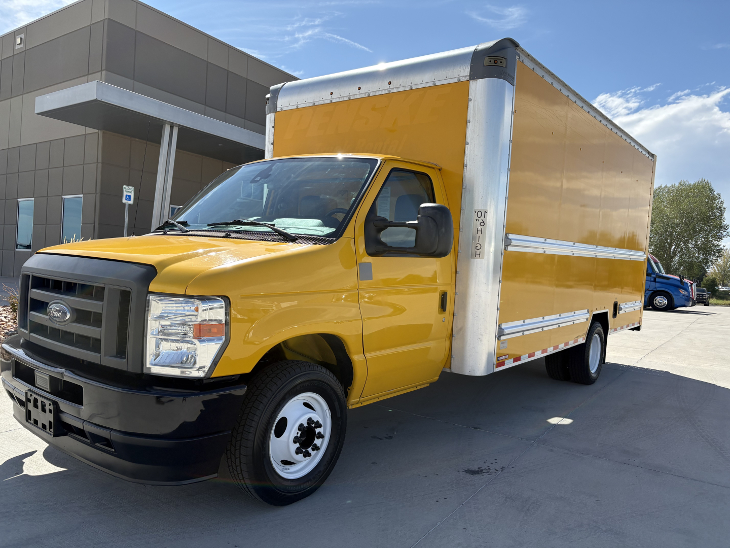 Yellow Ford E-350 box truck in a parking lot, front left view with cargo box and black grille, dealership building in background.
