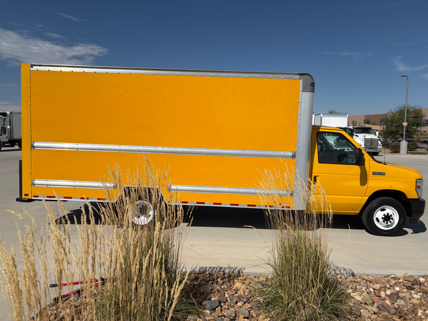 Side view of a yellow 2022 Ford E350 box truck with a large orange cargo box on a paved lot, wheel wells, and clear blue sky.