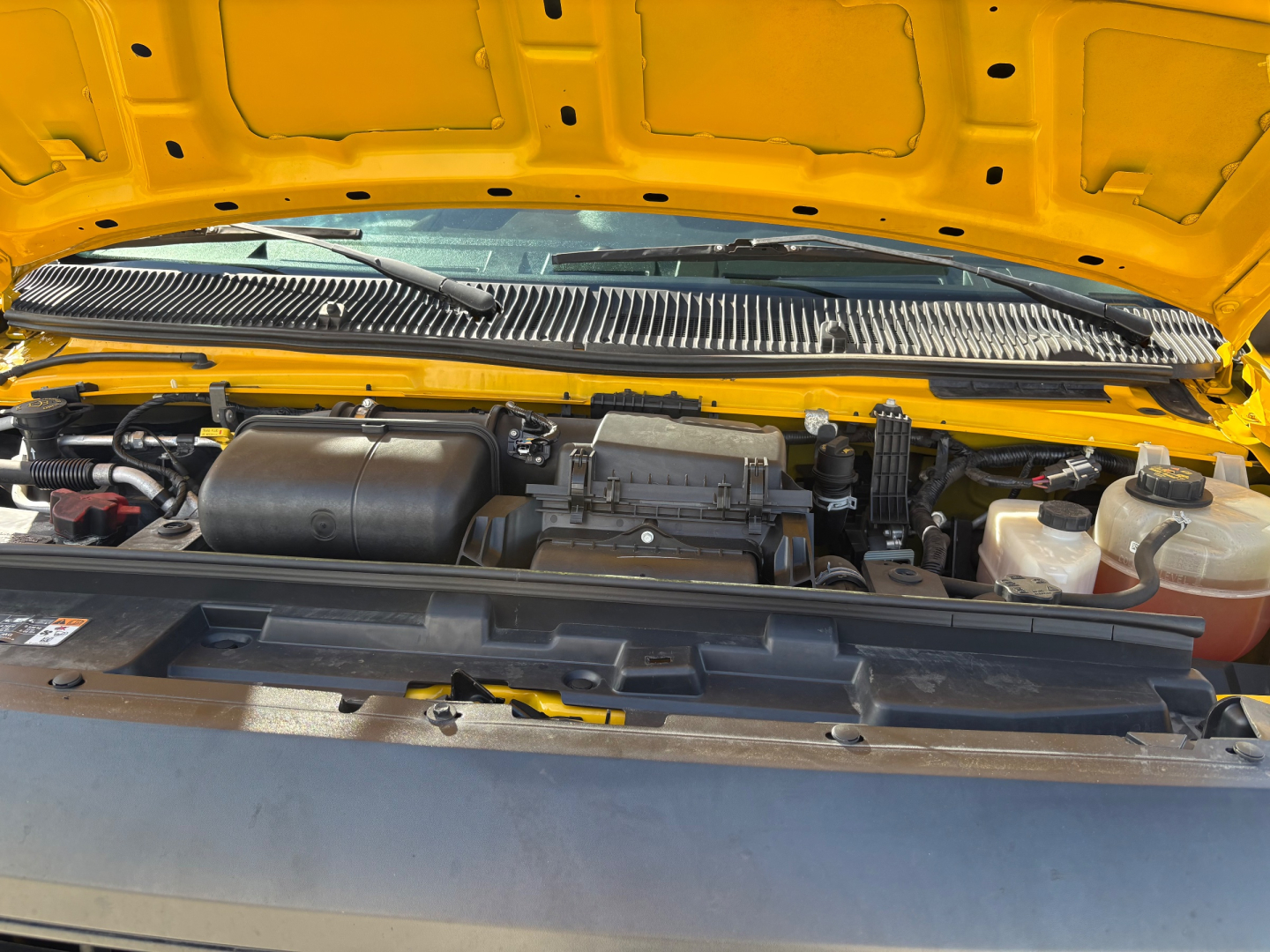 Ford E-350 engine bay under yellow hood showing air intake, battery, coolant reservoir, and various engine components.