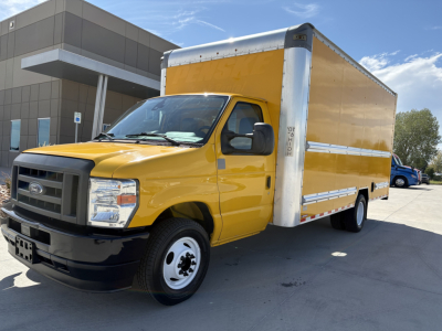 Yellow Ford E-350 box truck in a parking lot, front left view with cargo box and black grille, dealership building in background.