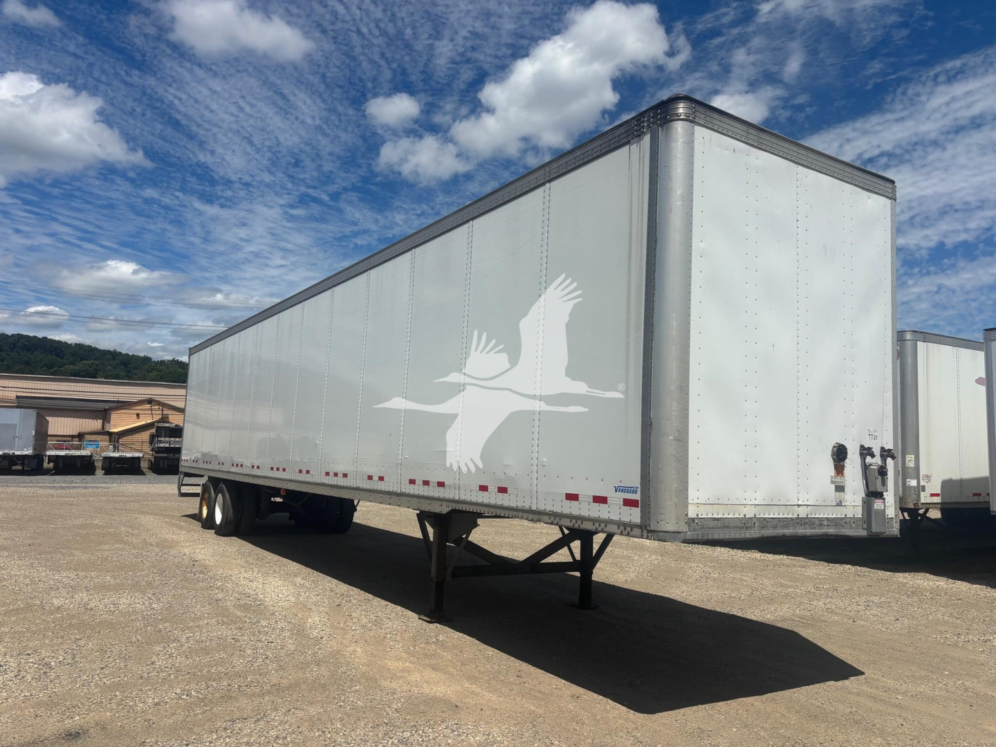 Trailer side view showing white Vanguard logo and profile of a bird on a silver box trailer under a blue sky with clouds.