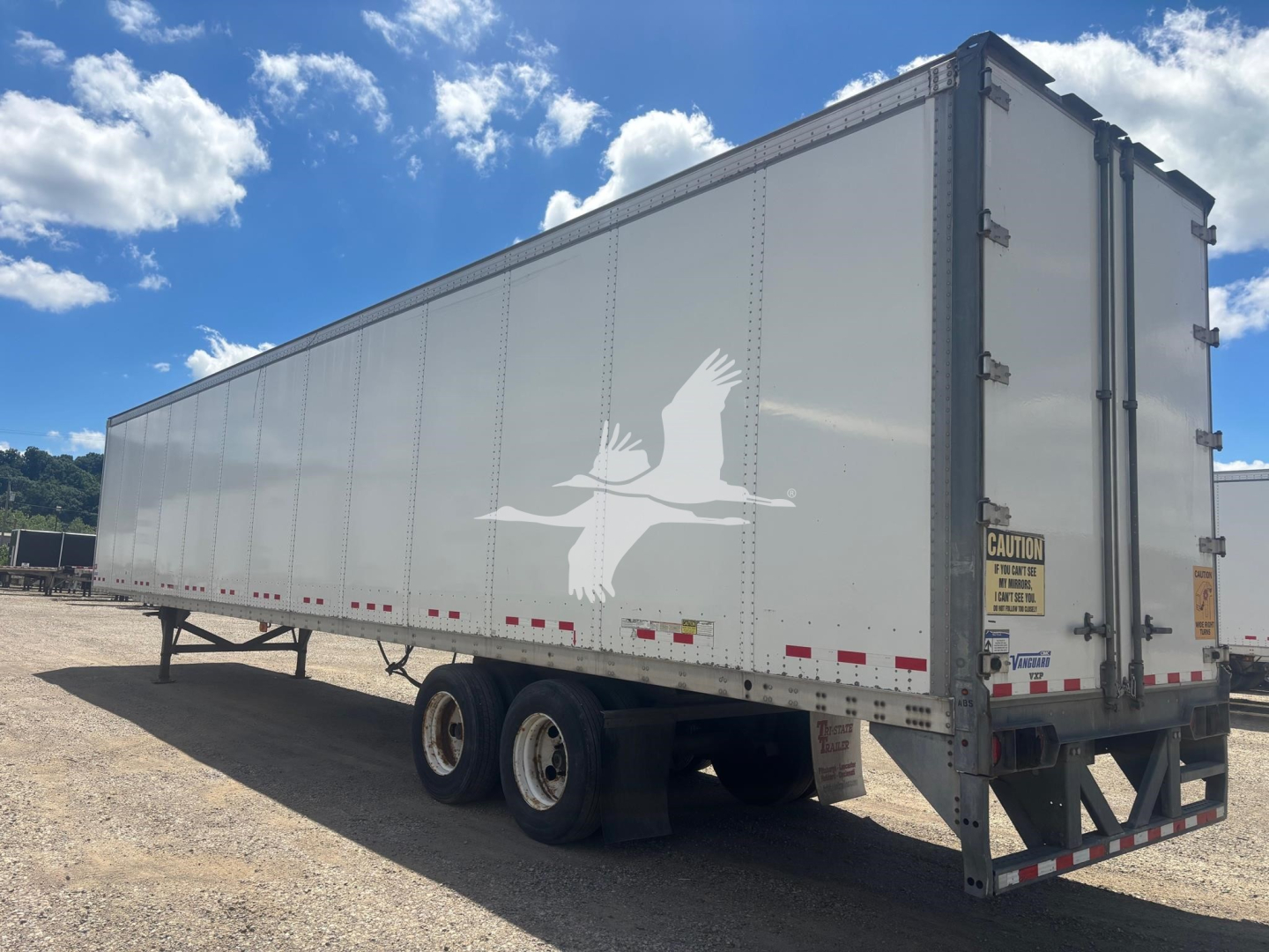 Vanguard trailer rear side view showing triple-axle undercarriage and CAUTION sticker on corner post