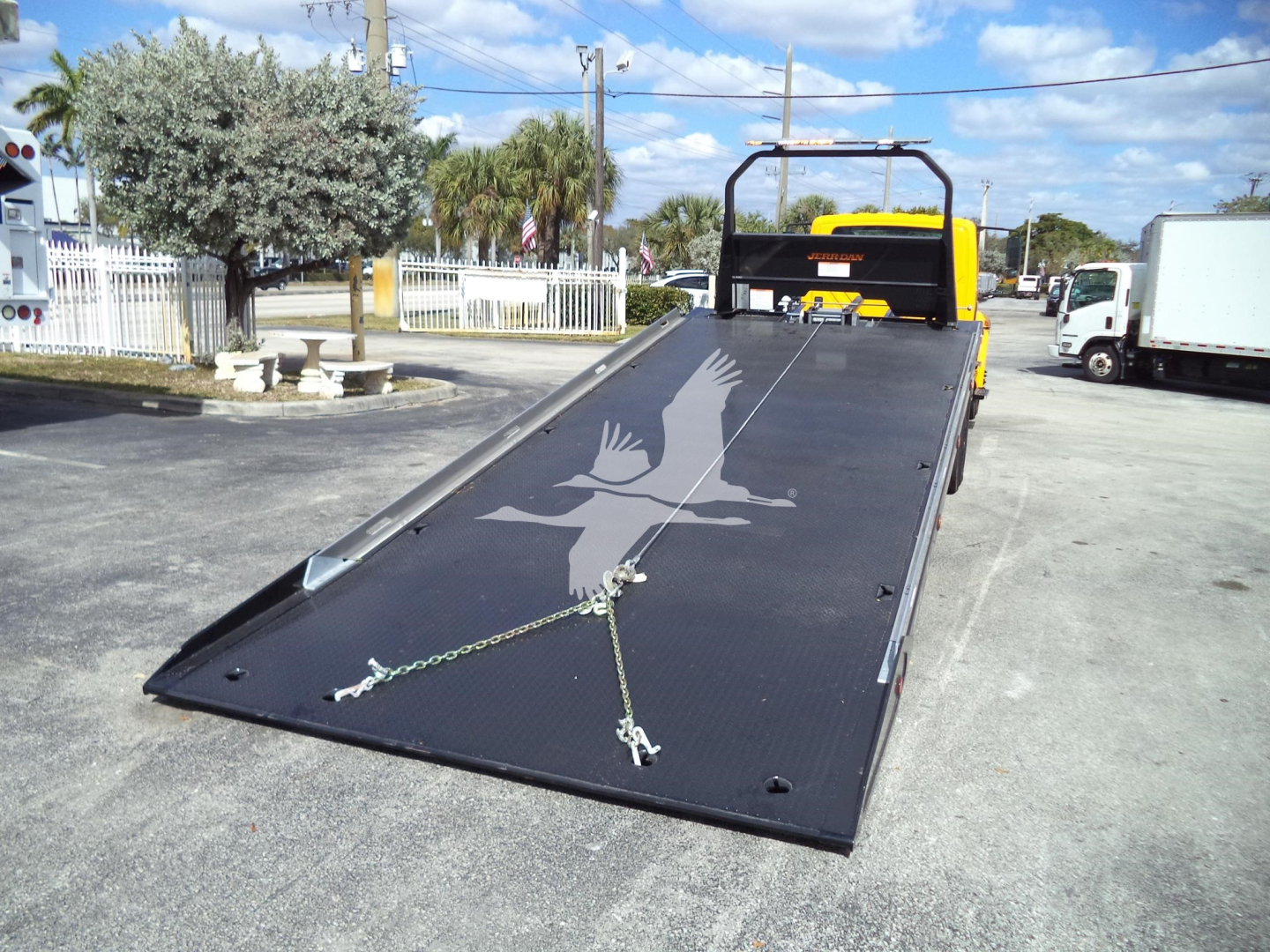 Flatbed tow truck with a ramp extended, black surface with a white eagle logo and chains on the ramp; truck cab visible in background.