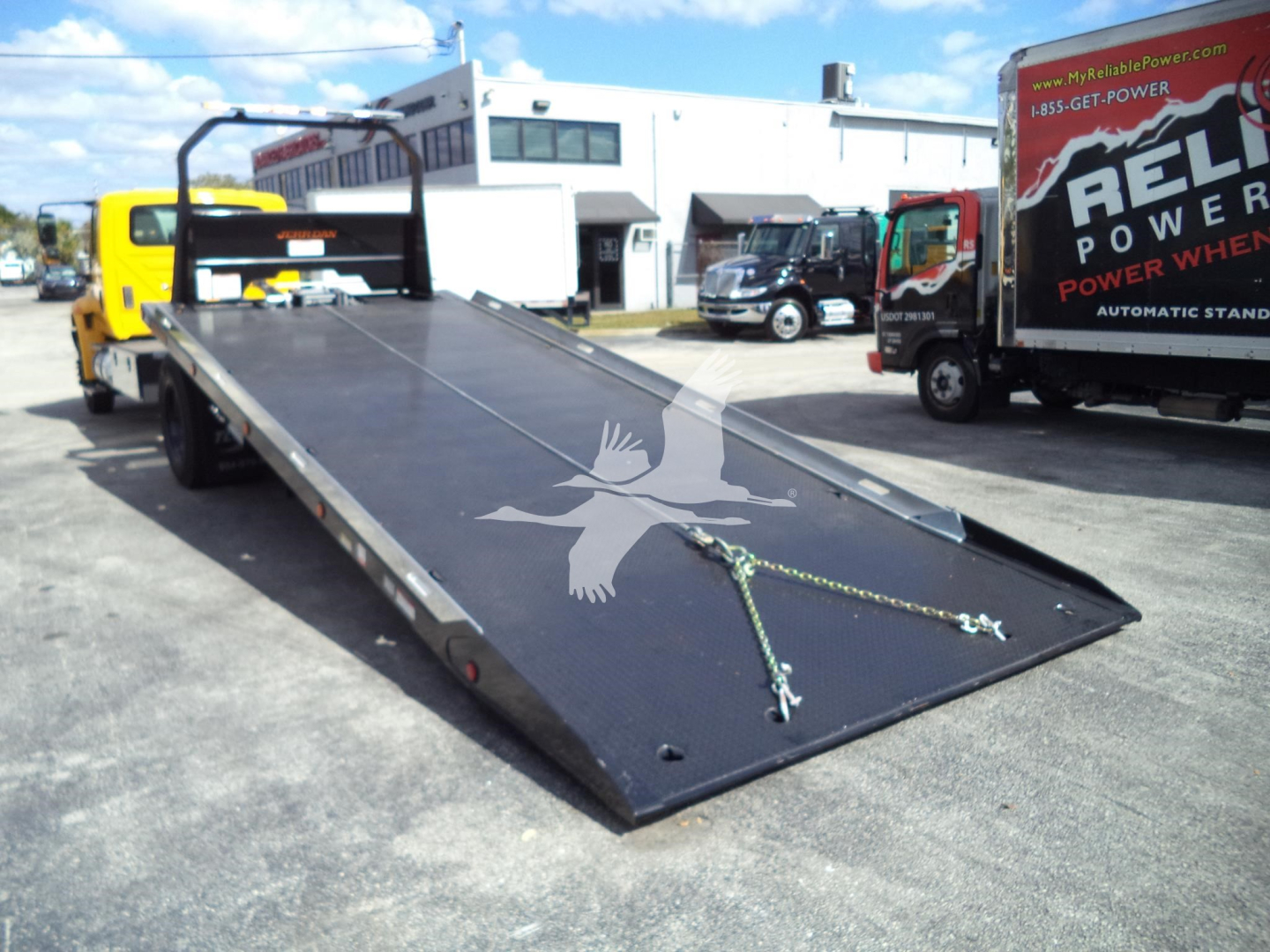 Flatbed tow truck with long black loading deck, lowered bed, and chains, in a dealership lot with other trucks in background.