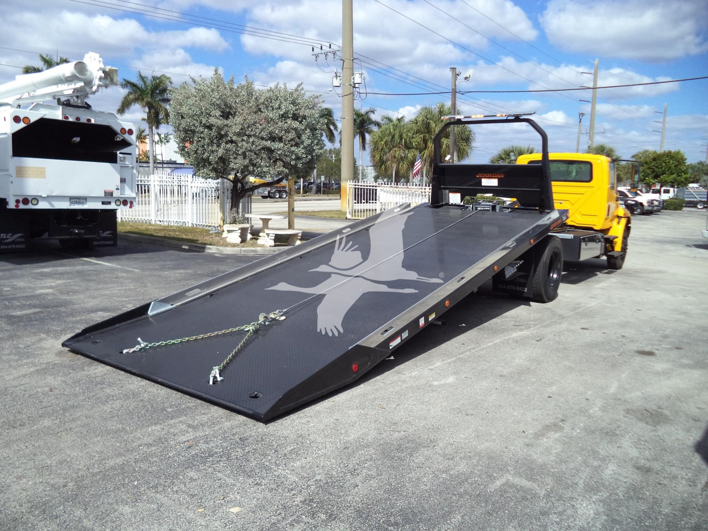 Flatbed tow truck with yellow cab and black ramp in lowered position; bird graphic on ramp and urban lot background.