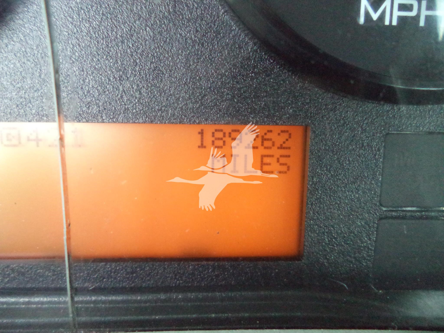 Dashboard instrument cluster showing speedometer with orange digital display and overlayed logo. Numbers and text illegible.
