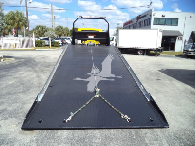 Truck wrecker flatbed on a 2019 International 4300 rollback showing a black steel deck with chains and hooks, yard with other trucks in background.