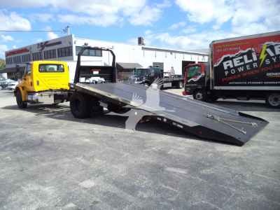 2019 International 4300 rollback wrecker flatbed on truck yard. Visible text includes REEL? POWER? on side of a truck in background, and a yellow tow truck attached to a long tilt-bed with black surface and side rails.