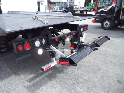 Truck bed with hydraulic rollback platform and folded steel ramp, multiple lights, and towing hooks visible at rear.