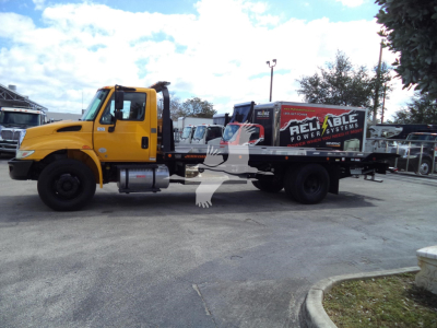 Vehicle side view of a yellow International 4300 rollback tow truck with a black flatbed, logoed with Reliable Powersystems on the bed.