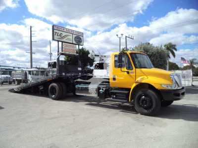 Truck image showing a yellow International 4300 rollback wrecker chassis with a black flatbed, side view, daytime.