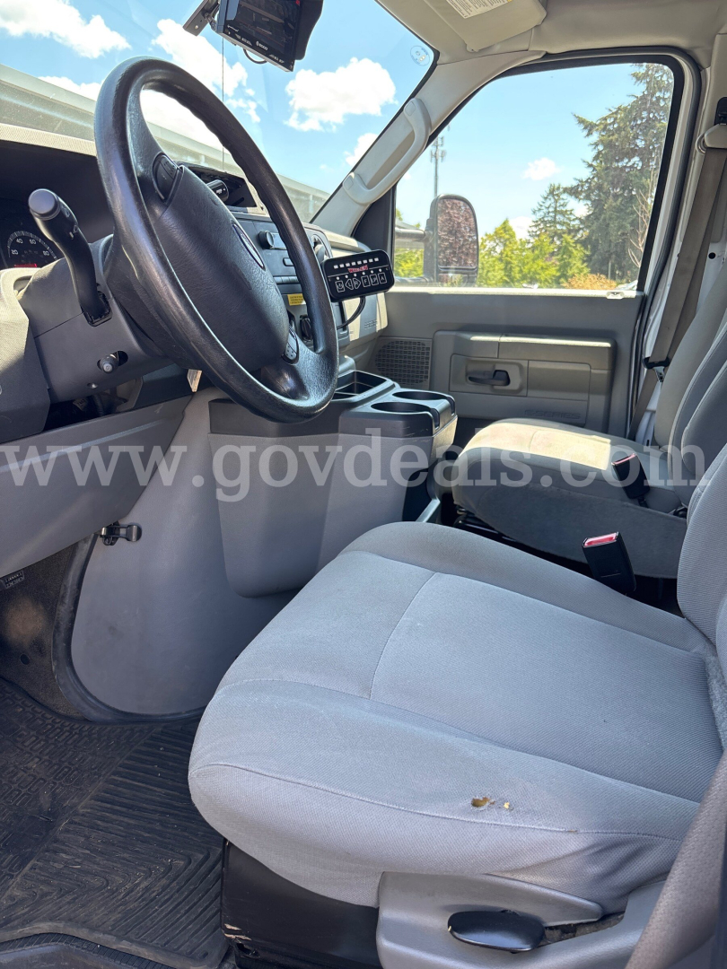 Front interior view of a 2017 Ford Econoline cab showing steering wheel, dashboard, and gray cloth driver seat with minor wear.