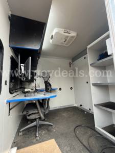 Ford Econoline interior showing workspace with computer, chair, storage shelves, and ceiling mounted device.