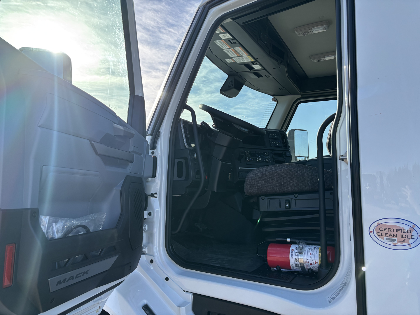 Cab interior of a new 2027 Mack AN64T daycab truck showing steering wheel, dashboard, and open driver door with a fire extinguisher on the floor.