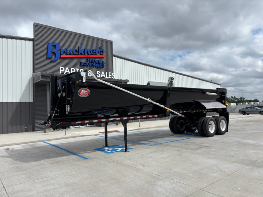 Trailer side view showing a black dump trailer with extendable gooseneck and dual rear axles parked in lot; Buckner’s Trucks sign in background.