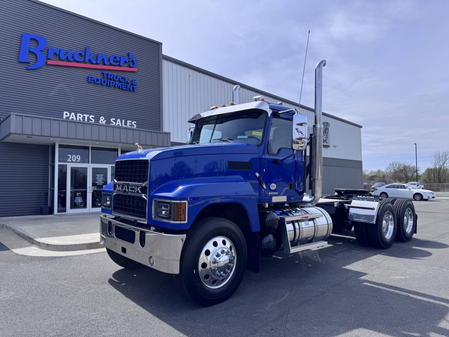Mack PI64T conventional daycab truck in blue parked outside a parts and sales building showing chrome grille, dual rear axles, and exhaust stack.