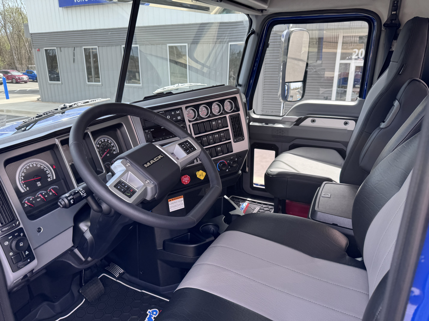 Mack PI64T truck interior cab view showing steering wheel, dashboard with gauges, and control panel. Text on dashboard includes Mack logo and various switches.