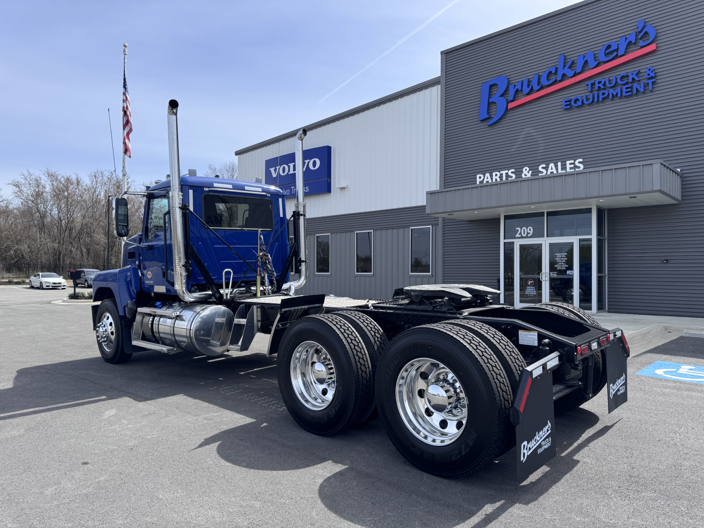 Truck chassis in blue cab-over-engine Mack PI64T with dual rear axles and fuel tanks, parked outside a parts and sales building