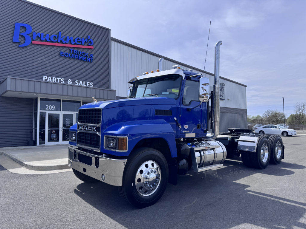 Mack PI64T conventional daycab truck in blue parked outside a parts and sales building showing chrome grille, dual rear axles, and exhaust stack.