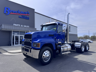 Mack PI64T conventional daycab truck in blue parked outside a parts and sales building showing chrome grille, dual rear axles, and exhaust stack.