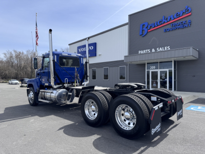 Truck chassis in blue cab-over-engine Mack PI64T with dual rear axles and fuel tanks, parked outside a parts and sales building