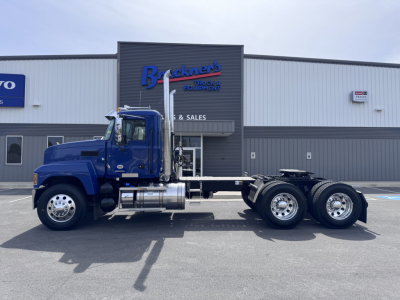 2027 Mack PI64T conventional daycab truck side view showing blue cab and chassis with triple rear axles in a dealership lot.