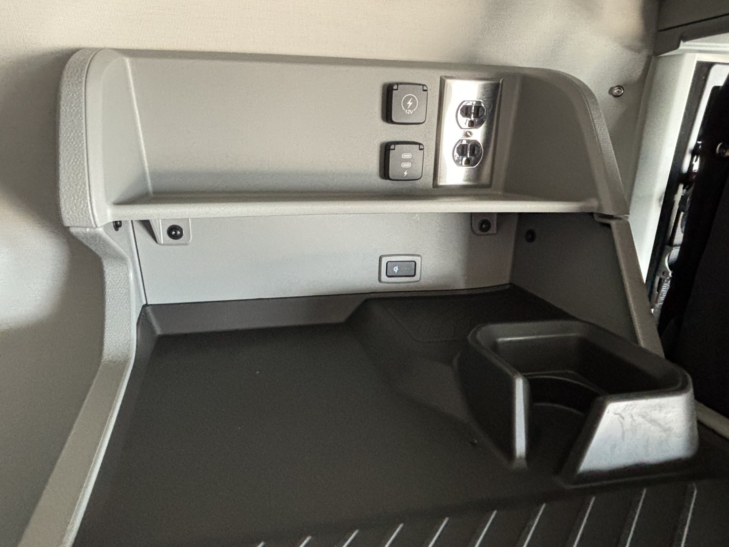 Cab area of a 2027 Volvo VNL sleeper showing control panel with two round ports and switches; grey storage shelf and cup holder.