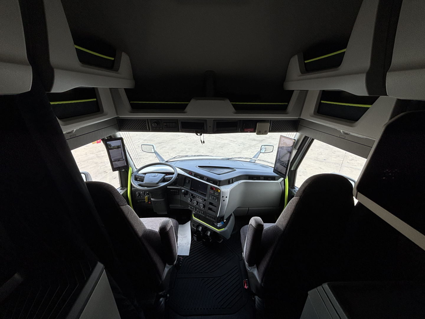 Interior view of a Volvo VNL64T860 cab. Front dashboard with steering wheel, large windshield, and overhead storage; two seats visible with side mirrors and driver’s monitor.