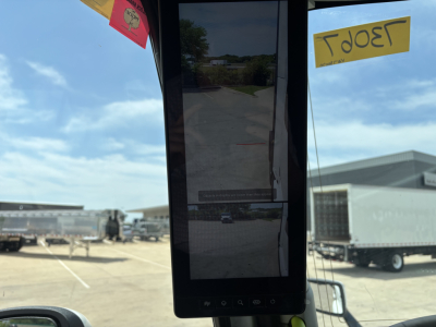 Truck cab interior rearview mirror area with tall vertical screen and windshield view; yellow price tags visible through glass.