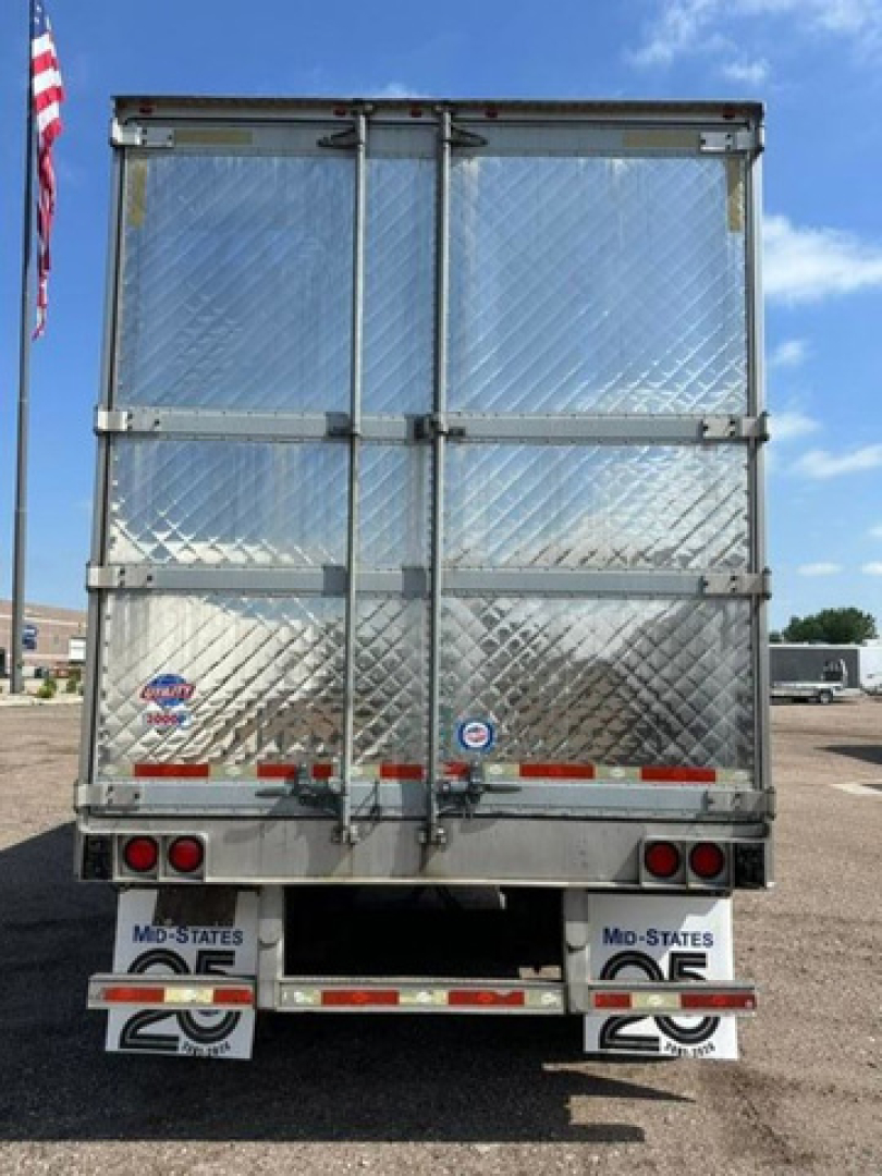 Rear view of a silver refrigerated trailer with diamond plate doors, barriers, and taillights. License/branding stickers near bottom, U.S. flag reflected in surface.