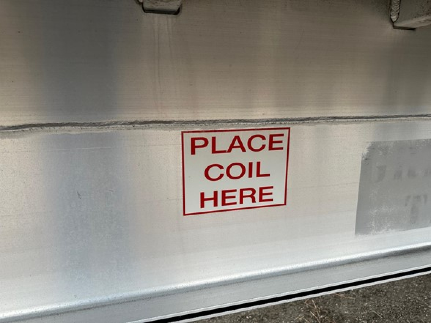 Flatbed trailer side showing a rectangular red-outlined sign with 'PLACE COIL HERE' stencil on metal surface.